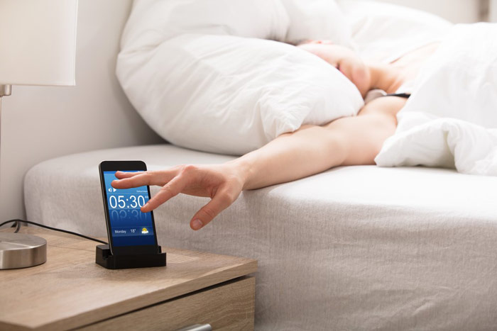 Człowiek mający problemy z porannym wstawaniem – wyciąga spod kołdry rękę, by wyłączyć alarm w telefonie - zdrowy-sen.eu