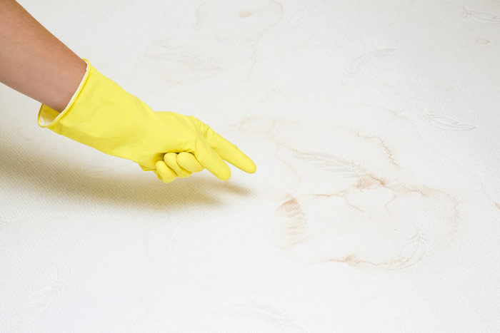 Dłoń w żółtej, gumowej rękawiczce ochronnej wskazująca na plamę na materacu - zdrowy-sen.eu