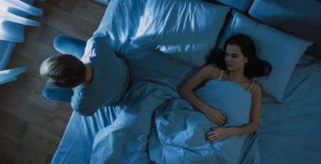 Mężczyzna, który nie może zasnąć w stresie, siedzi w nocy na łóżku w sypialni obok leżącej na tym samym łóżku kobiety - zdrowy-sen.eu