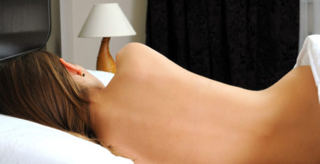 Spanie nago – młoda kobieta leżąca w swoim łóżku, przykryta do połowy, pokazana od tyłu - zdrowy-sen.eu