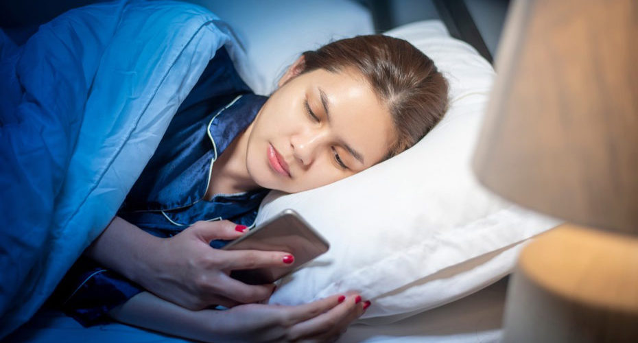 Kobieta mająca trudności ze snem, leżąca w łóżku z telefonem komórkowym w ręku - zdrowy-sen.eu